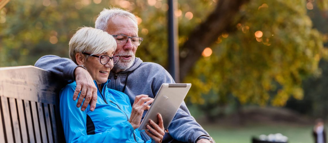 Smiling senior active couple sitting on the bench looking at tab